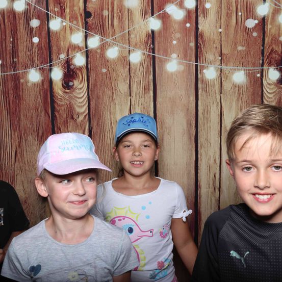 Vier Kinder posieren vor einem hölzernen Hintergrund mit Lichterketten. Das Mädchen in der Mitte trägt eine blaue Kappe mit dem Namen Sophie. Das Mädchen auf der linken Seite trägt eine rosa Kappe mit dem Wort Hello. Das Mädchen auf der rechten Seite trägt ein weißes T-Shirt mit einem Seepferdchen-Design. Der Junge auf der rechten Seite lächelt.