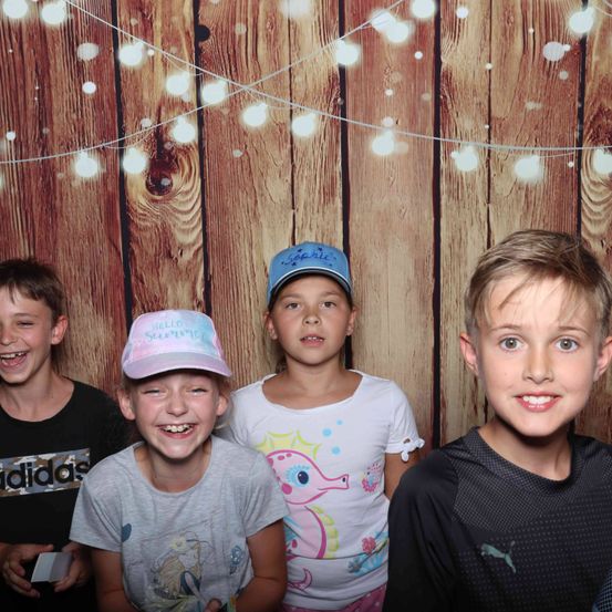 Vier Kinder lächeln in einem Fotoautomaten mit hölzerne Hintergrund und Lichterkette. Zwei Mädchen und zwei Jungen, einer trägt eine Baseball-Kappe, posieren zusammen.