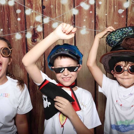 Drei Kinder in weißen T-Shirts posieren für ein Foto vor einem hölzernen Hintergrund mit einer Piraten-Mütze, Schutzbrille und einem Piraten-Bandana.