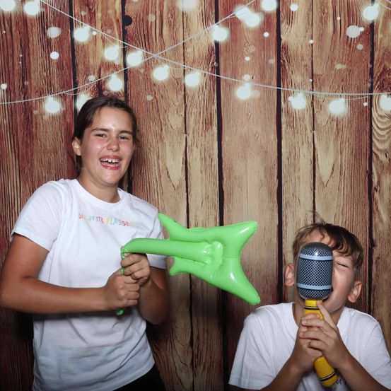Zwei Kinder in weißen T-Shirts spielen mit einer grünen Ballongitarre und einem Mikrofon. Sie lächeln und posieren für ein Foto vor einem hölzernen Hintergrund mit weißen Lichtern.