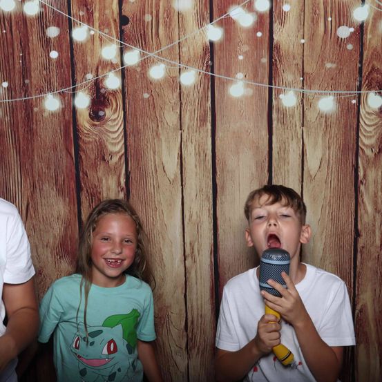 Ein junger Junge singt in ein Mikrofon, während ein Mädchen und eine weitere Person in der Nähe stehen. Sie stehen vor einem hölzernen Hintergrund mit dekorativen Lichtern.