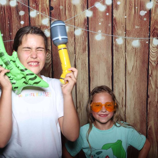 Zwei Mädchen posieren für ein Foto mit einer grünen aufblasbaren Gitarre und einem gelben Mikrofon vor einer Holzwand. Das Mädchen auf der linken Seite trägt ein weißes T-Shirt mit bunter Schrift. Das Mädchen auf der rechten Seite trägt orange Sonnenbrille.