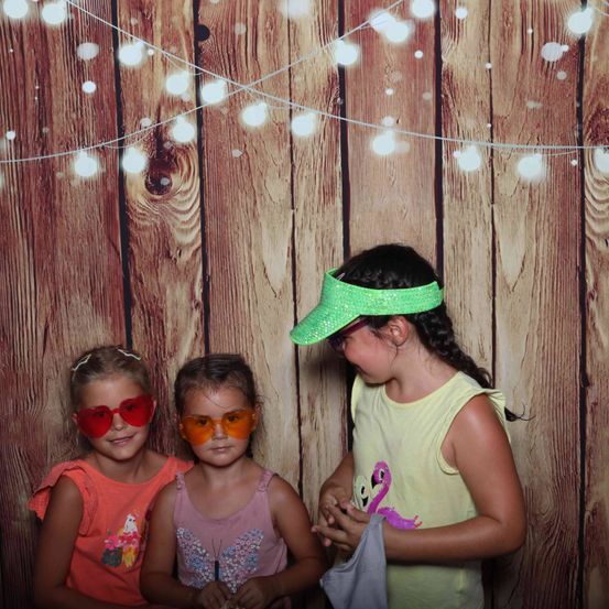 Drei junge Mädchen posieren für ein Foto vor einer Holzwand mit Lichtern. Sie tragen Sonnenbrillen und ein Mädchen hat eine grüne Schirmmütze.