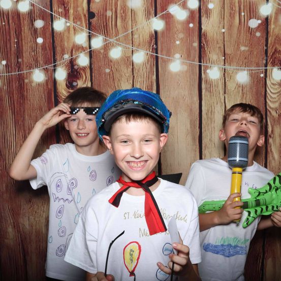 Drei Jungen posieren vor einem hölzernen Hintergrund mit Lichtern. Der Junge in der Mitte trägt einen blauen Hut und einen roten Schal. Der Junge auf der linken Seite hält eine Sonnenbrille und der Junge auf der rechten Seite hält ein Mikrofon und eine Spielzeuggitarre.