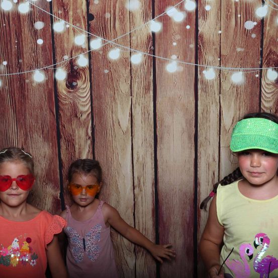 Drei junge Mädchen posieren für ein Foto vor einem mit Lichterketten geschmückten hölzernen Hintergrund. Sie tragen Sonnenbrillen und leuchtend farbenfrohe Shirts.