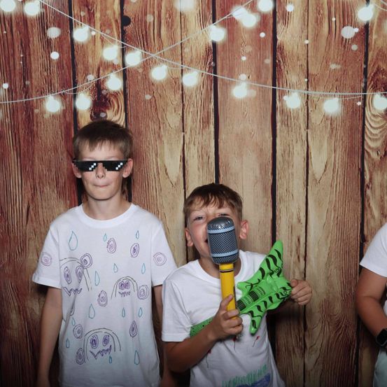 Zwei Jungen stehen vor einer Holzwand mit Lichterketten. Ein Junge trägt ein weißes T-Shirt mit bunten Zeichnungen, und der andere Junge hält ein Mikrofon und eine Spielzeuggitarre.