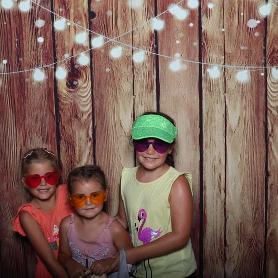 Drei junge Mädchen mit Sonnenbrillen und lächelnd, stehen vor einem hölzernen Hintergrund mit Lichterketten.