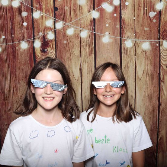 Zwei Mädchen tragen pixelierte Brillen und lächeln vor einem hölzernen Hintergrund mit dekorativen Lichtern. Ihre weißen T-Shirts haben handgezeichnete Wolken und Phrasen wie 'Die beste' und 'Klasse'.