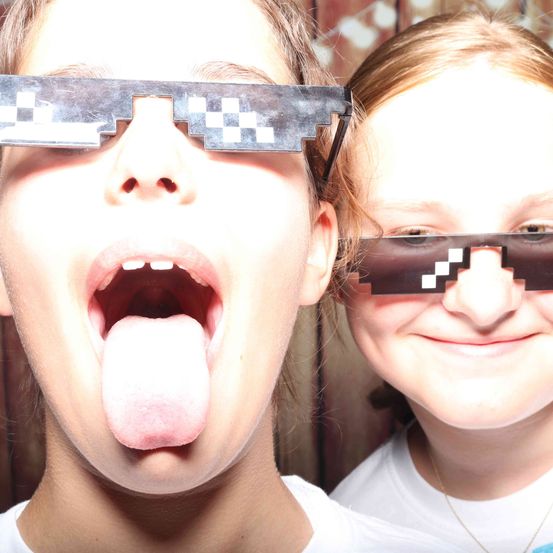 Zwei junge Menschen tragen Minecraft-Stil-Brillen, einer schiebt die Zunge heraus. Sie stehen nah beieinander und lächeln für die Kamera.