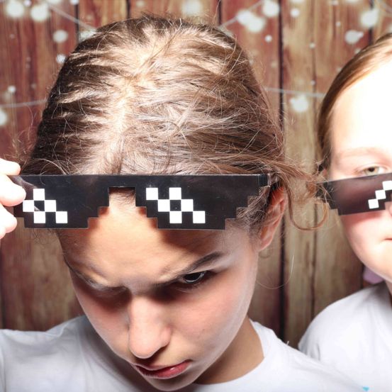 Zwei Mädchen mit Minecraft-Themenbrillen posieren für ein Foto vor einem hölzernen Hintergrund. Das Mädchen vorne sieht ernst aus.
