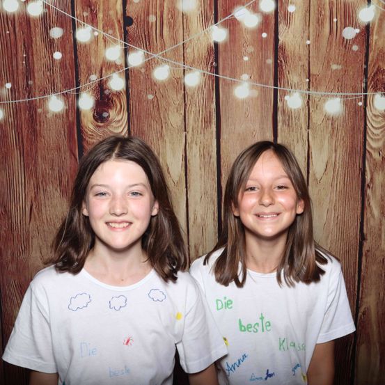 Zwei junge Mädchen stehen gemeinsam vor einem hölzernen Hintergrund mit Lichtern. Sie tragen weiße T-Shirts mit handgeschriebenen Botschaften. Das Mädchen links hat 'Die beste' auf ihrem Shirt stehen, während das Mädchen rechts 'Anna' und 'beste Klasse' hat. Beide lächeln.
