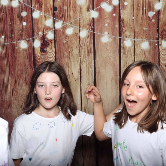 Zwei Mädchen posieren fröhlich für ein Foto vor einem hölzernen Hintergrund mit Lichterketten. Sie tragen weiße T-Shirts mit bunten Zeichnungen und Text.