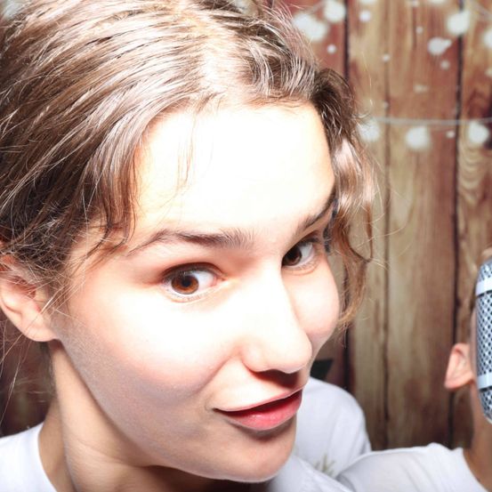 Eine Frau mit kurzen, welligen braunen Haaren schaut direkt in die Kamera. Sie hat braune Augen und trägt ein weißes T-Shirt. Hinter ihr ist der Kopf einer Person mit einem Mikrofon zu sehen. Der Hintergrund ist eine Holzwand.