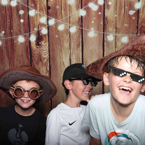 Drei Jungen in Hüten und Sonnenbrillen posieren für ein Foto vor einem hölzernen Hintergrund mit Lichterketten.