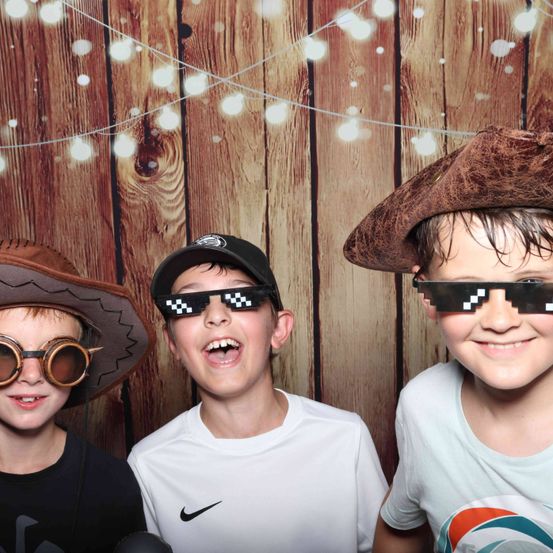 Drei Jungen stehen vor einem hölzernen Hintergrund mit Lichterketten. Einer trägt eine Brille, ein anderer eine Kappe und der dritte einen Piratenhut. Sie alle lächeln für die Kamera.