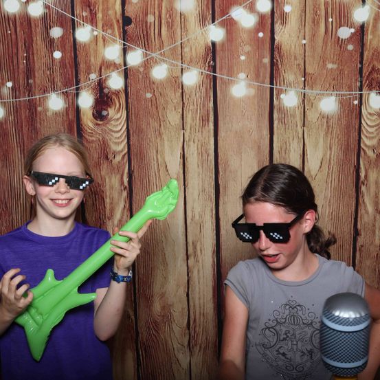 Zwei junge Mädchen posieren für ein Foto, eine hält eine neongrüne Gitarre und die andere ein Mikrofon. Sie lächeln und tragen pixelierte Sonnenbrillen. Ein hölzerner Hintergrund mit Lichterketten sorgt für eine spaßige Atmosphäre.