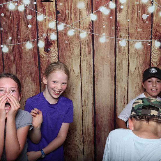 Vier Kinder stehen vor einer Holzwand mit Lichterketten. Das Mädchen auf der linken Seite hat die Hände im Gesicht. Der Junge in der Mitte trägt ein lila Hemd. Der Junge rechts trägt eine Tarnkappe.