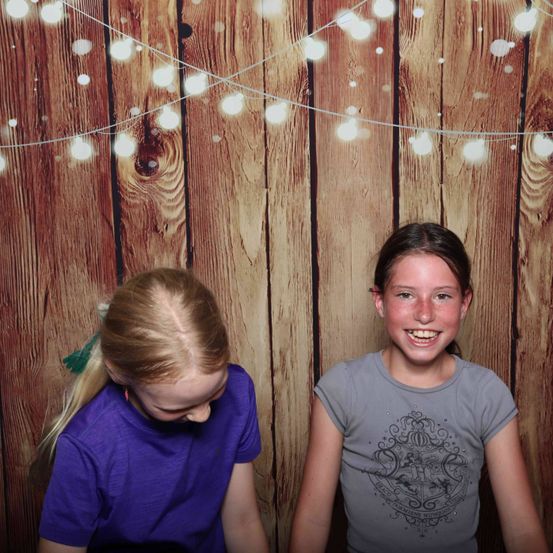 Zwei Mädchen posieren für ein Foto vor einem hölzernen Hintergrund mit weißen Lichtern. Das Mädchen auf der linken Seite trägt ein lila T-Shirt, während das Mädchen auf der rechten Seite ein graues T-Shirt mit Grafikdesign trägt.