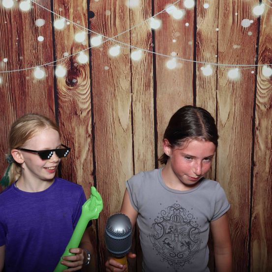 Zwei junge Mädchen stehen vor einem mit Lichterketten geschmückten Holzhintergrund. Das Mädchen auf der linken Seite trägt Lila und hält eine grüne Handpuppe, während das Mädchen auf der rechten Seite Grau trägt und ein Mikrofon hält. Beide scheinen für ein Foto zu posieren.