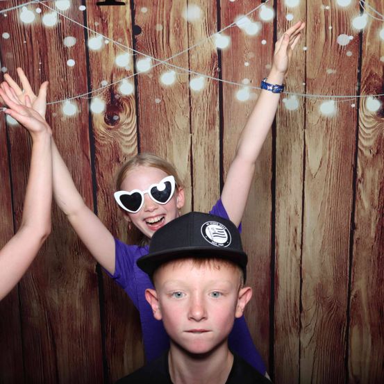 Drei Kinder sind in einer Fotokabine. Das Mädchen trägt herzförmige Sonnenbrillen und ein Armband. Der Junge trägt einen schwarzen Hut. Der hölzerne Hintergrund hat weiße Lichter.