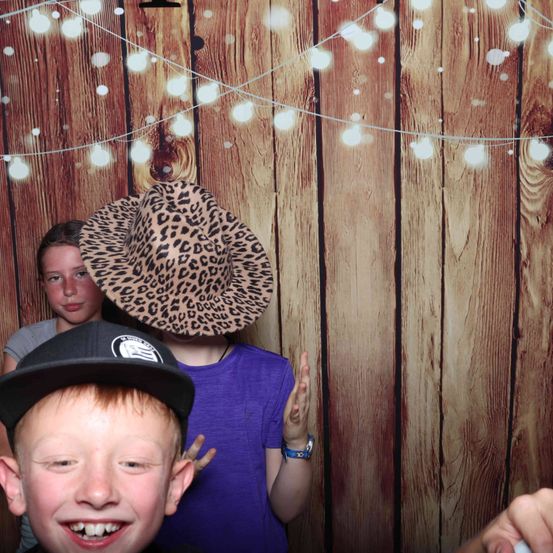 Drei Kinder posieren vor einem hölzernen Hintergrund mit Lichtern. Einer trägt einen Leoparden-Print Hut, ein anderer ein lila T-Shirt und der dritte einen schwarzen Hut.