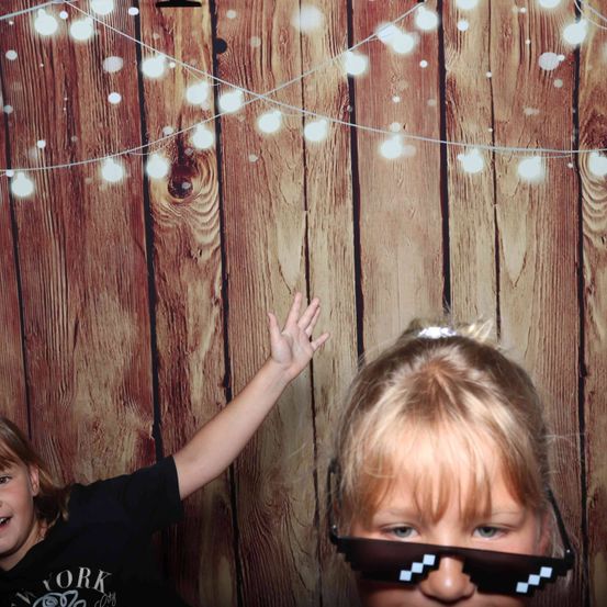 Zwei Kinder stehen vor einer Holzwand mit Lichterketten. Das Mädchen trägt Minecraft-Brillen. Der Junge hebt seine Hand.