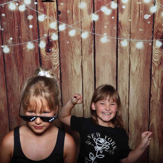 Zwei junge Mädchen posieren für ein Foto vor einem hölzernen Hintergrund mit Lichterketten, eines trägt Pixelbrille und das andere ein schwarzes New York T-Shirt.