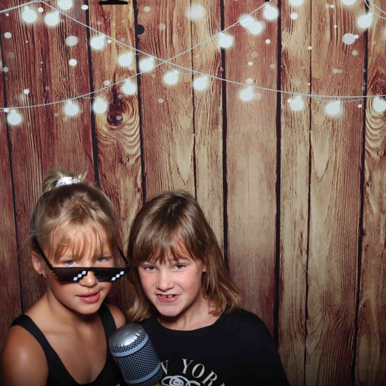 Zwei junge Mädchen stehen gemeinsam vor einer Holzwand mit Lichterketten. Das Mädchen auf der linken Seite trägt pixelierte Brille und ein schwarzes Tanktop. Das Mädchen auf der rechten Seite trägt ein schwarzes T-Shirt mit dem Wort 'New York'. Ein Mikrofon ist zwischen ihnen positioniert.