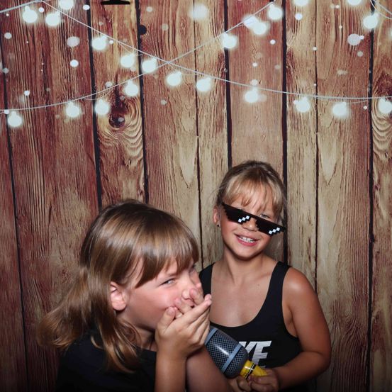 Zwei junge Mädchen posieren für ein Foto vor einem hölzernen Hintergrund mit Lichterketten. Ein Mädchen trägt eine Sonnenbrille und hält ein Mikrofon, während das andere Mädchen ihren Mund mit der Hand bedeckt.
