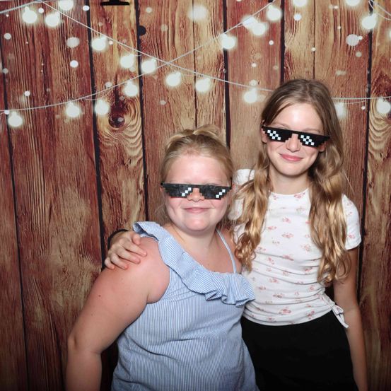 Zwei junge Mädchen mit pixeligen Sonnenbrillen posieren gemeinsam vor einem Holzhintergrund mit dekorativen Lichtern.