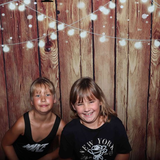 Zwei Kinder, ein Junge und ein Mädchen, lächeln und posieren für ein Foto vor einem hölzernen Hintergrund mit Lichterketten.