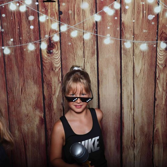 Ein junges Mädchen mit einem schwarzen Tanktop mit Nike-Logo und pixeligen Sonnenbrillen posiert vor einem hölzernen Hintergrund mit Lichterketten.
