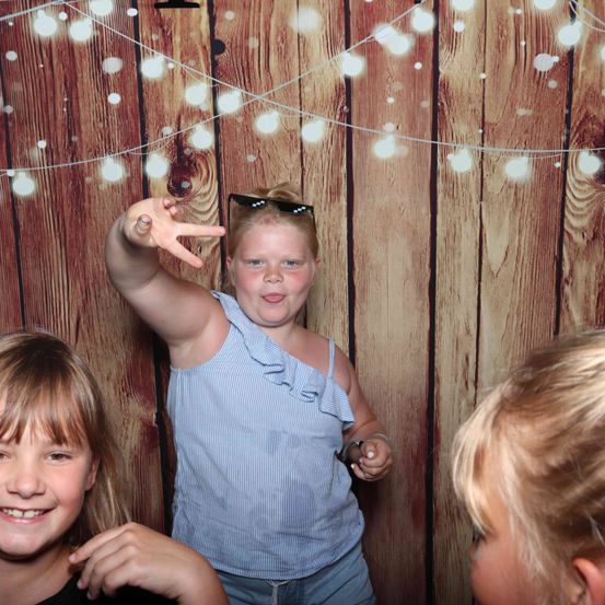 Drei junge Mädchen stehen vor einem hölzernen Hintergrund. Das mittlere Mädchen macht ein Friedenszeichen.