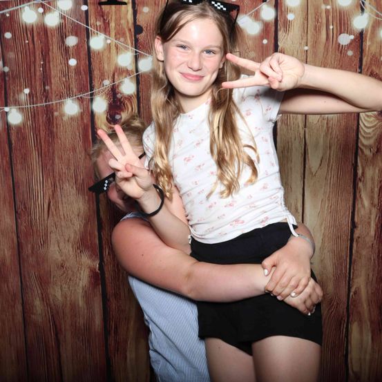 Ein Mädchen mit einem weißen Shirt mit Blumendesigns posiert mit einem Friedenszeichen-Handgeste, während es von einer anderen Person vor einer Holzwand umarmt wird.