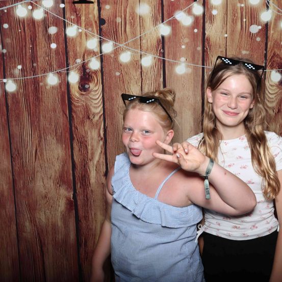 Zwei junge Mädchen mit Brillen auf dem Kopf posieren für ein Foto vor einer Holzwand mit Lichterketten.