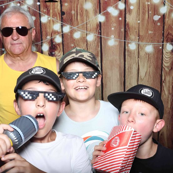 Vier Jungen posieren für ein Foto. Drei tragen Sonnenbrillen, einer hält ein Mikrofon und ein anderer eine Popcorntüte. Hinter ihnen steht ein älterer Mann in der Nähe einer Holzwand mit Lichtern.