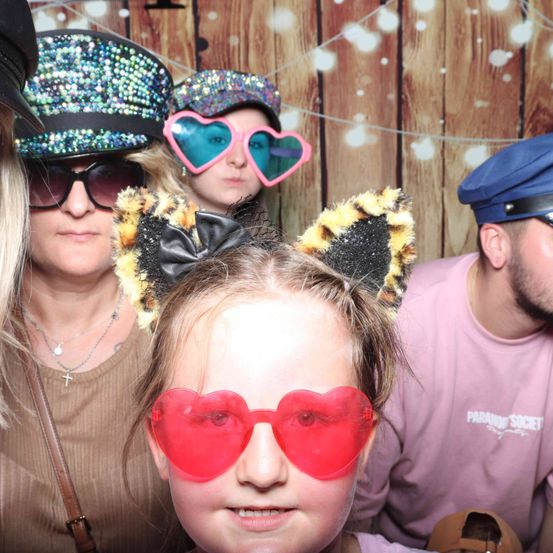 Vier Personen posieren für ein Foto vor einem hölzernen Hintergrund. Ein Mädchen trägt herzförmige Brillen und Katzenohren. Zwei Frauen tragen funkelnde Hüte und ein Mann eine dunkelblaue Mütze.