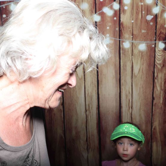 Eine ältere Frau mit weißen Haaren lächelt ein kleines Mädchen an, das eine grüne Schirmmütze trägt, in einem Fotokabine mit Holz-Thema.