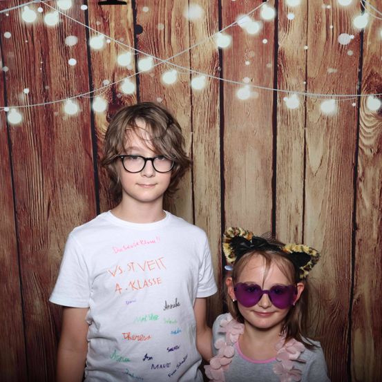 Zwei Kinder stehen vor einem hölzernen Hintergrund mit Lichtern. Ein Junge mit Brille und ein Mädchen mit Katzenohren lächeln in die Kamera. Der Junge trägt ein T-Shirt mit bunter Beschriftung.