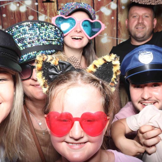 Eine Gruppe von Menschen mit Hüten und Brillen posiert für ein Foto vor einem hölzernen Hintergrund. Ein Mädchen trägt Katzenohren und andere tragen herzförmige Brillen.