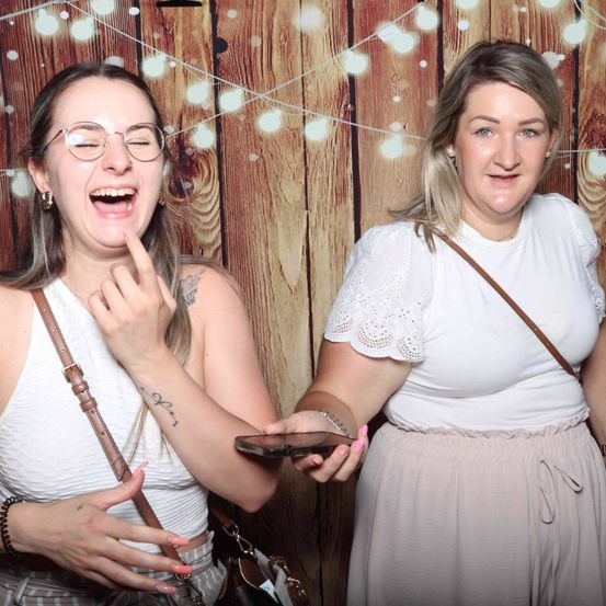 Zwei Frauen posieren für ein Foto in einer Fotobox mit einem hölzernen Hintergrund. Eine Frau trägt eine Brille und ein Tattoo, während die andere ein Telefon hält.