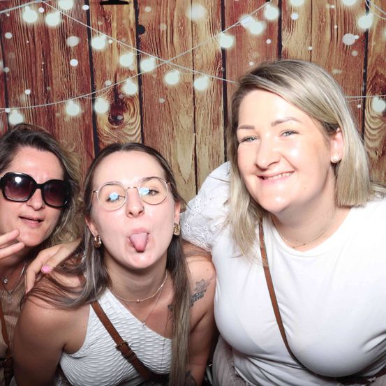Drei Frauen posieren für ein Foto vor einem hölzernen Hintergrund. Die Frau auf der linken Seite trägt eine Sonnenbrille. Die mittlere Frau sticht ihre Zunge heraus. Die Frau auf der rechten Seite lächelt. Sie tragen Halsketten und weiße Oberteile.