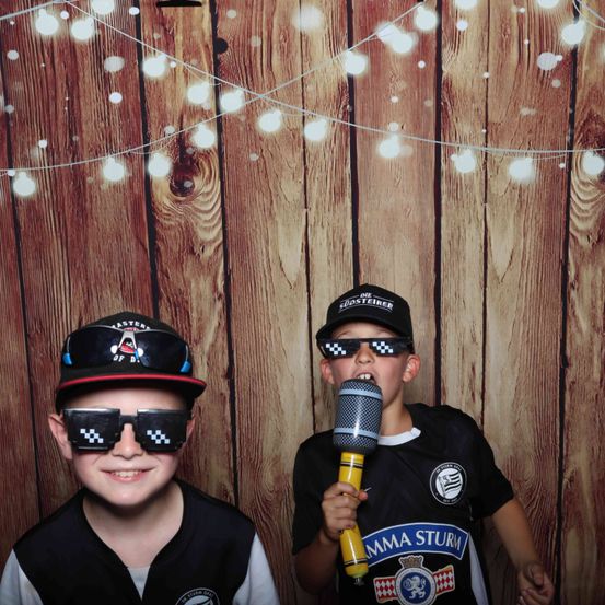 Zwei Jungen in einem Fotostudio, die schwarze Westen, Mützen und Sonnenbrillen tragen. Einer hält ein Mikrofon. Ein hölzerner Hintergrund mit Lichterketten.