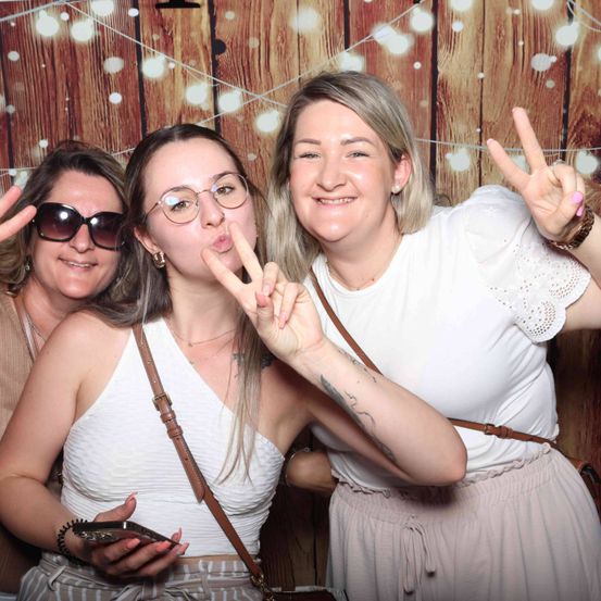 Drei Frauen posieren für ein Foto vor einem hölzernen Hintergrund und machen Friedenszeichen. Eine Frau trägt eine Brille und hält ein Telefon. Die anderen tragen weiße Oberteile und haben Tattoos.