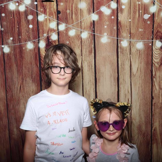 Zwei Kinder stehen vor einer Holzwand mit Lichtern. Ein Junge trägt eine Brille und ein weißes T-Shirt mit Aufschrift. Ein Mädchen trägt Katzenohr-Sonnenbrille und einen rosa Schal.