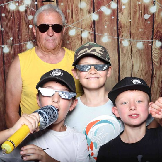 Drei Jungen und ein älterer Mann posieren für ein Foto im Studio. Der Mann trägt ein gelbes ärmelloses Shirt und die Jungen tragen Baseballkappen und Brillen. Ein Junge hält ein Mikrofon.