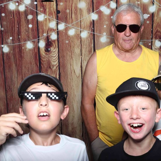 Ein älterer Mann steht hinter zwei Jungen, einer in Sonnenbrille und der andere in einer schwarzen Kappe, und posiert für ein Foto vor einem hölzernen Hintergrund mit Lichterketten.