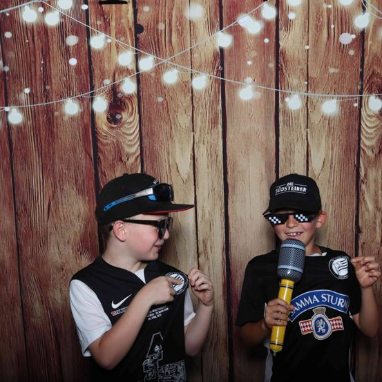 Zwei Jungen posieren vor einem hölzernen Hintergrund für ein Foto. Einer hält ein Mikrofon und trägt eine Kappe mit dem Text 'Gamma Sturm'. Der andere trägt eine Baseballkappe und Sonnenbrille.