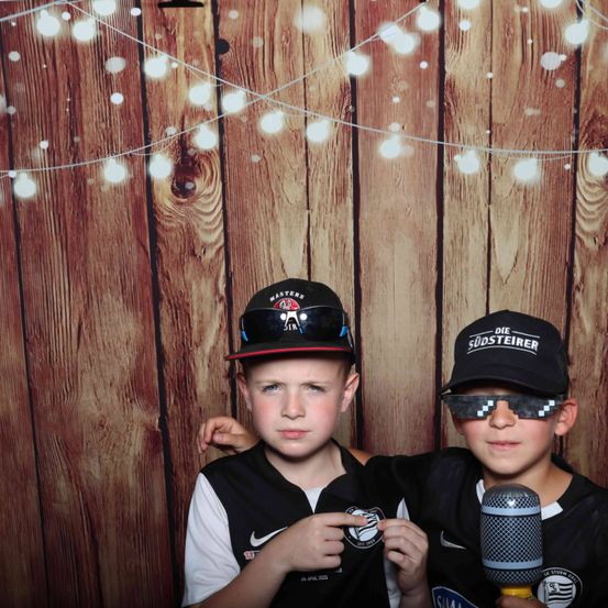 Zwei Jungen mit Baseballkappen und Mikrofonen posieren für ein Foto vor einer Holzwand mit Lichterketten.