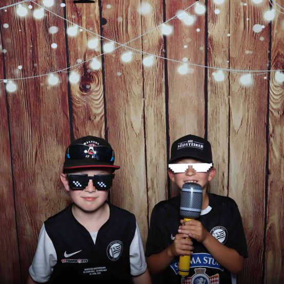 Zwei Jungen in Kostümen posieren für ein Foto vor einem hölzernen Hintergrund mit Lichterketten. Einer hält ein Mikrofon und der andere trägt eine Sonnenbrille.